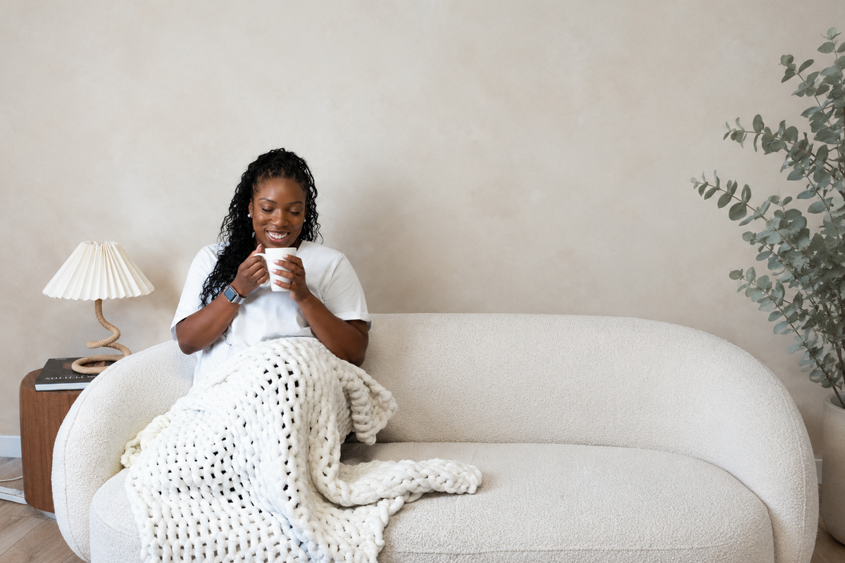 The founder of sante renata an intentional living wellness brand sits on a cream-colored sofa in a cozy living room setting, wrapped in a chunky white knit blanket and holding a white mug. A small wooden side table with a lamp is on the left, and a potted plant is on the right against a textured beige wall.