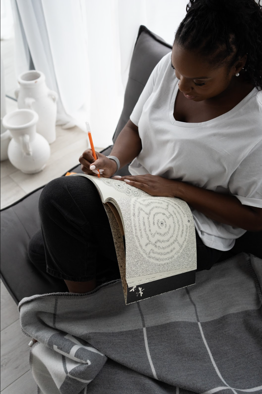 The founder of sante renata an intentional living brand thats launching wellness candles is sitting on a grey sofa, colouring in a colouring book with an orange pen. The page shows a spiral maze-like drawing.