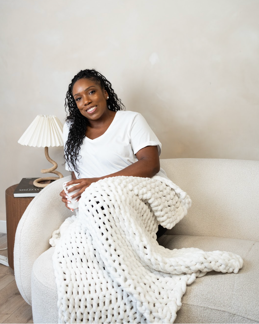 The founder of sante renata an intentional living wellness brand that sells wellbeing essential oil candles is sitting on a cream-colored sofa in a cozy living room setting, wrapped in a chunky white knit blanket. She is wearing a white t-shirt and holding a white mug.