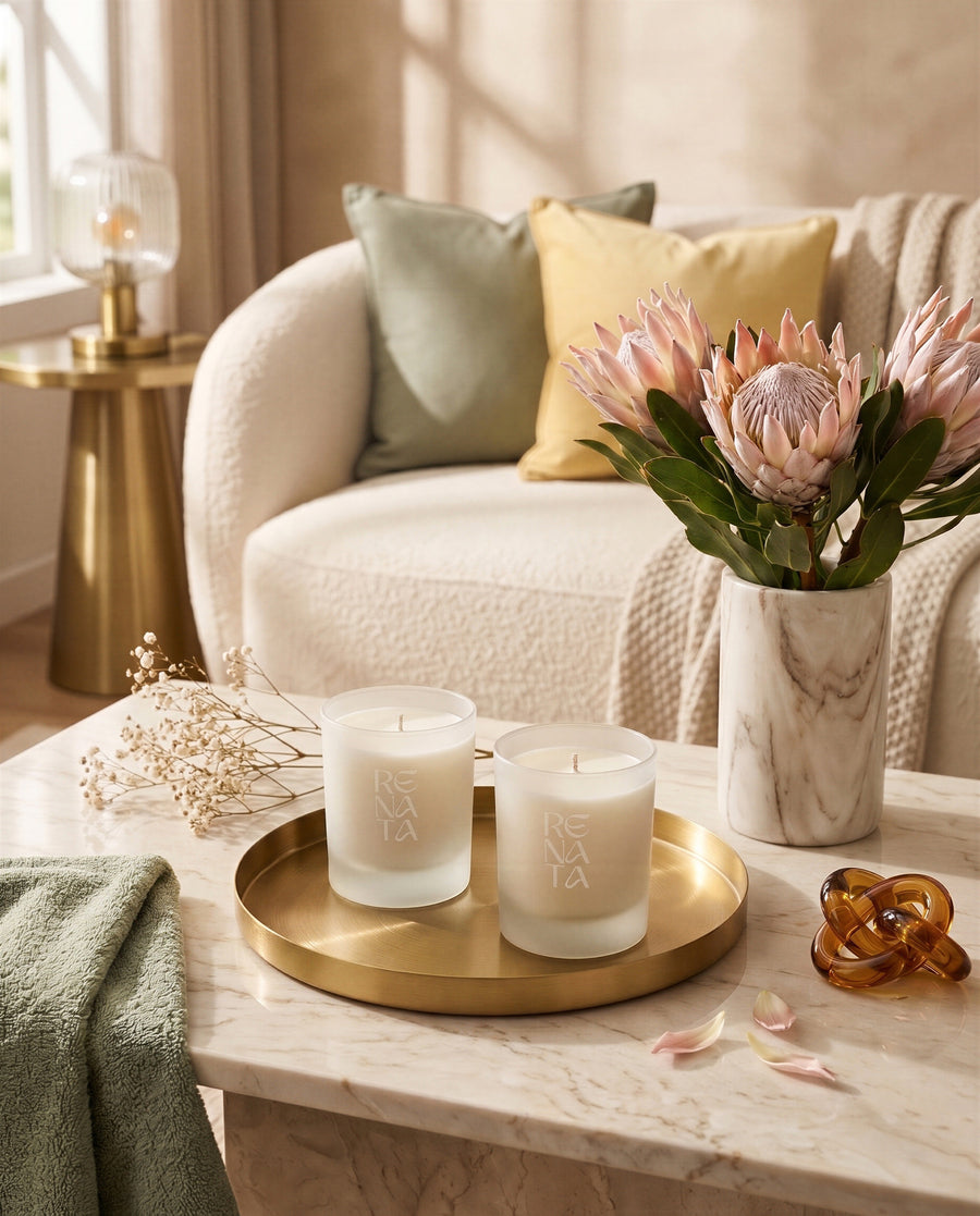 Two frosted glass candle jars with white wax and a minimalist logo, displayed on a gold tray on a marble coffee table in a cozy living room setting.