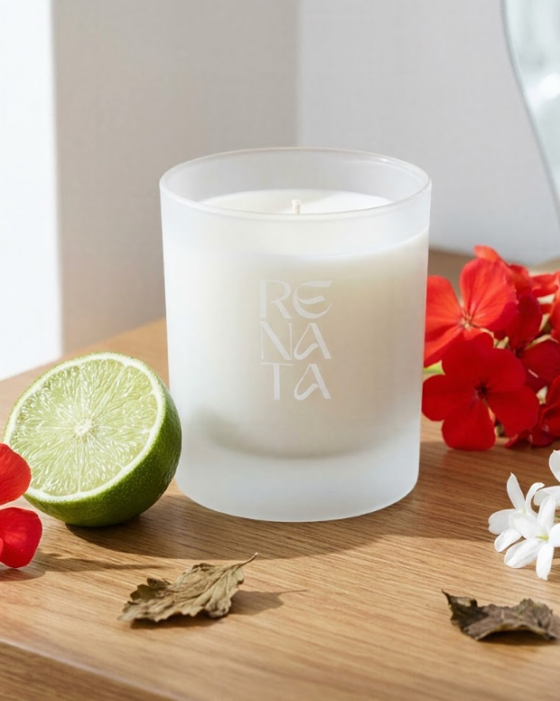 A frosted glass candle jar with white wax sits on a wooden surface, surrounded by red flowers, a lime half, and dried leaves. The candle is branded with the word 'RENTA' etched vertically on the front.